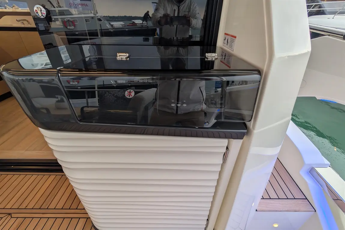 A sleek boat interior features a glossy black control panel and textured white cabinetry, with a view of water and boats outside.